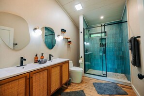 Un baño con lavamanos blanco, tocador de madera, dos espejos, inodoro y ducha con mampara de vidrio.
