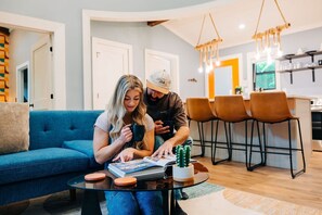 A modern living room with a blue sofa, a coffee table, and a dining area with chairs and a kitchen in the background. - Hot Tub + SxS Trails | Vibrant Silo Cabin | Kings (Beattyville)
