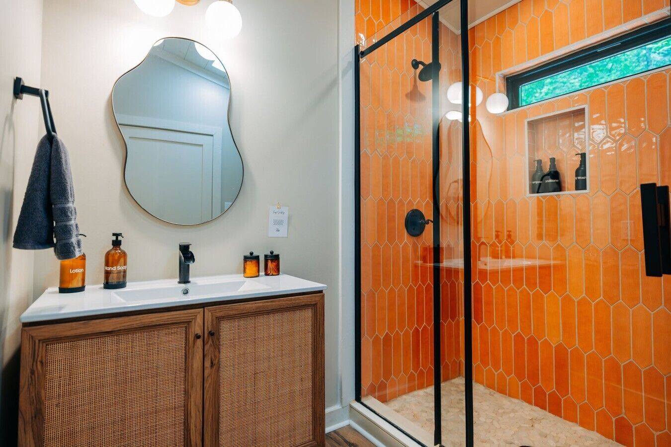 Een badkamer met een houten wastafelmeubel, een ronde spiegel en een douche met glazen wanden en oranje tegels.