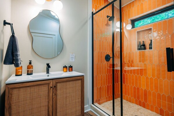 A bathroom with a wooden vanity, a round mirror, and a glass-enclosed shower with orange tiles.