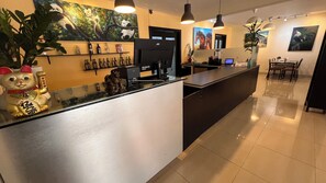 A modern hotel reception area with a counter, a JVC TV, and various decorations including a cat statue and artwork on the walls.