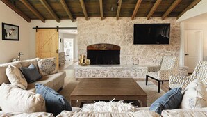 Una sala de estar con chimenea de piedra, vigas de madera en el techo, un sofá, un sillón estampado y un televisor de pantalla plana montado en la pared.