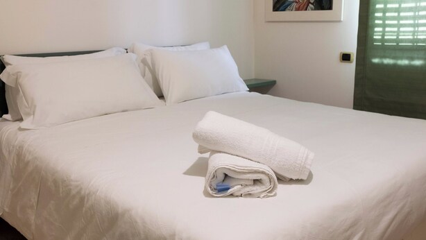 Una cama bien hecha con sábanas blancas y una toalla doblada, una mesita de noche con una lámpara, y un cuadro en la pared.