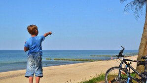 Escena de playa con un niño señalando al mar, una bicicleta estacionada cerca y un cielo despejado.