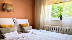 Una cama bien hecha con ropa de cama blanca y beige, una lámpara de pared tejida y una ventana con vistas a zonas verdes.
