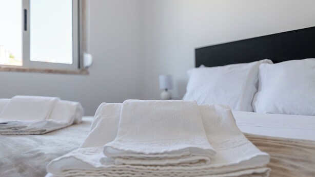 A neatly made bed with white linens and pillows, a bedside lamp, and a window with blinds. - Great remodeled apartment - 5 min to the beach! (Carcavelos)