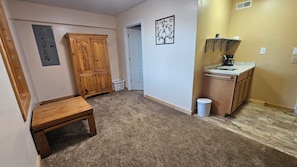 A hallway with a wooden bench, a white door, a small trash bin, and a kitchenette area with a sink and a coffee maker.