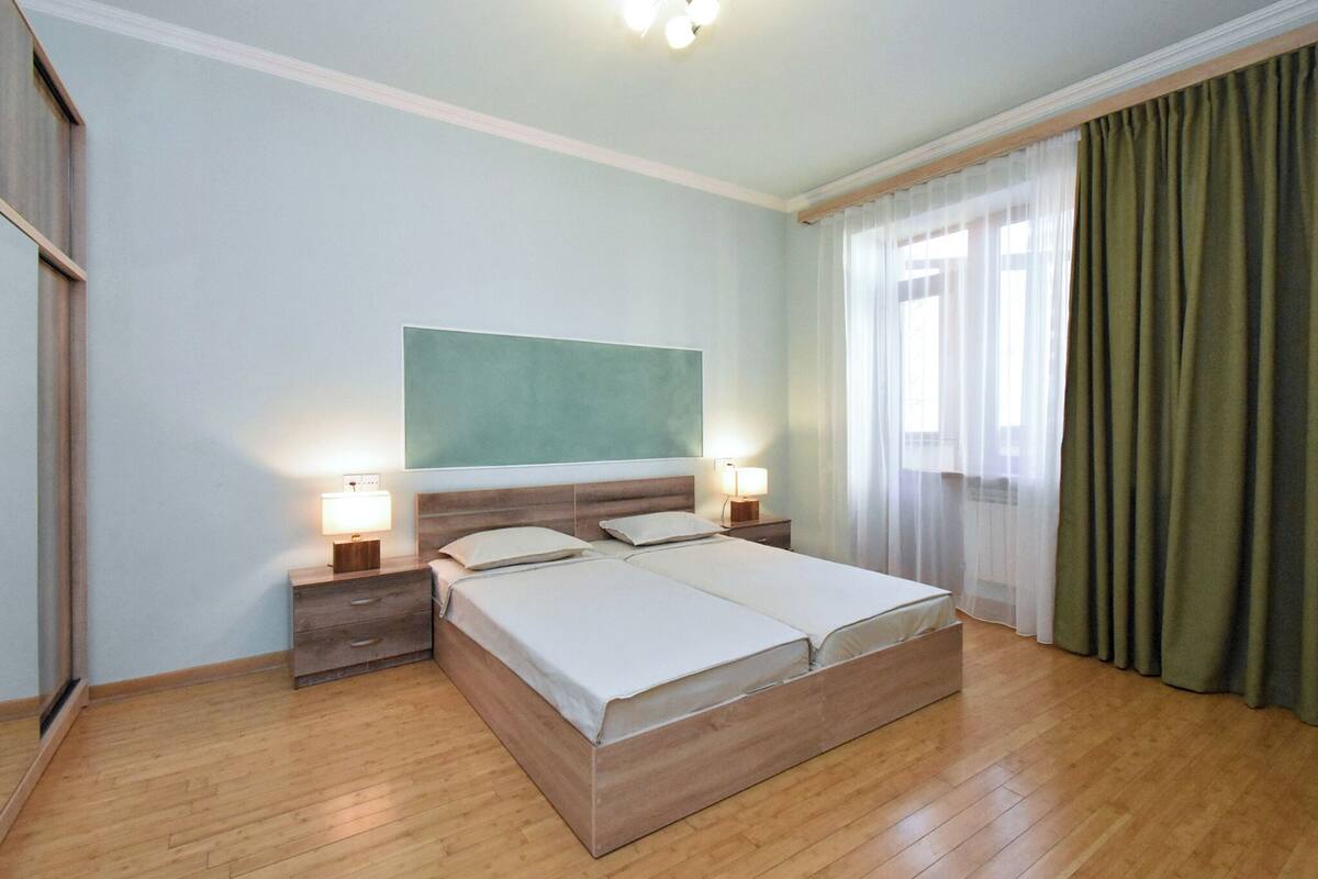 Cama doble con dos mesitas de noche, una mesita auxiliar, un cuadro abstracto grande en la pared y una ventana con cortinas traslúcidas.