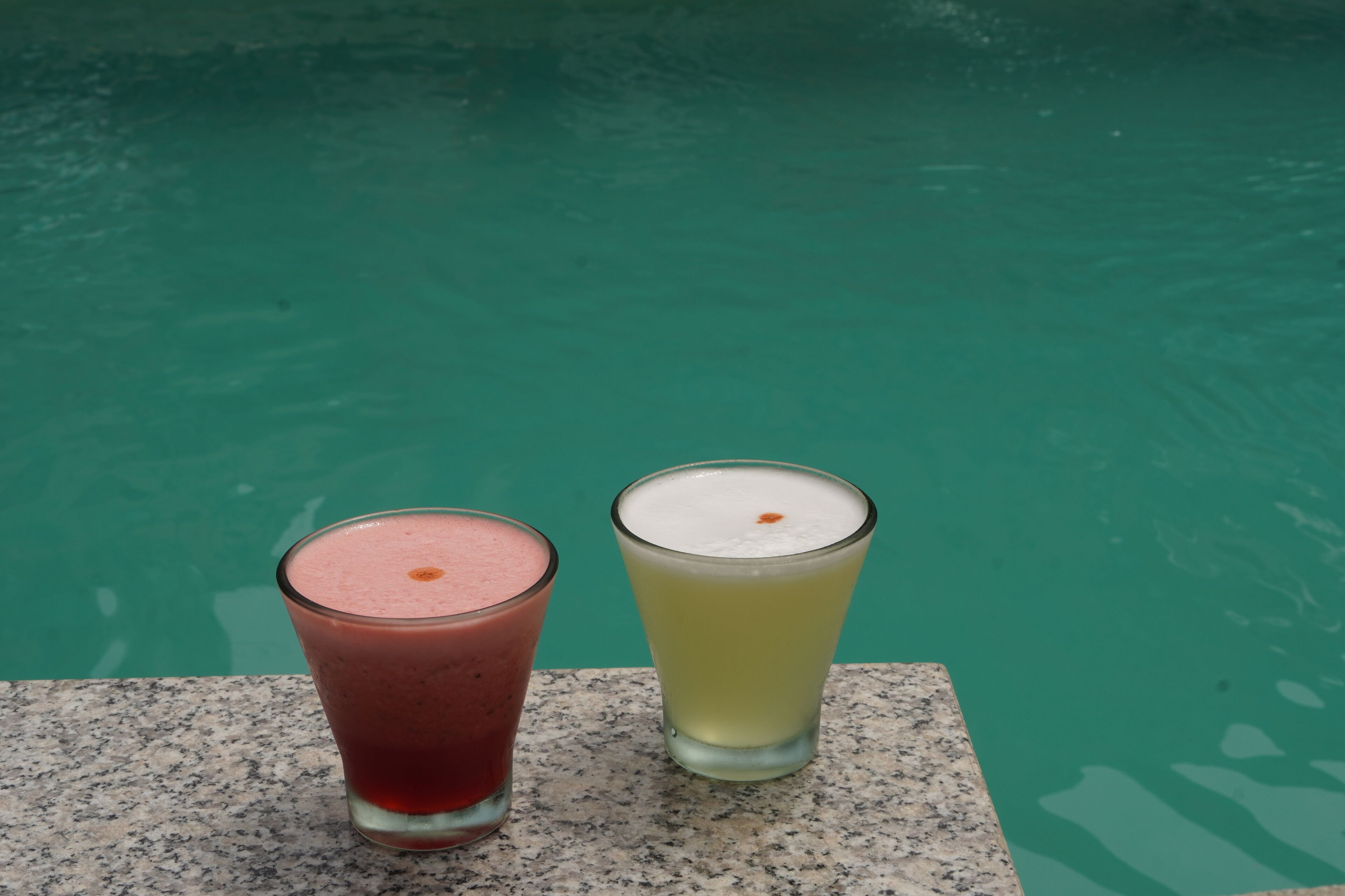 Deux verres de boissons sur une surface en granit, près d’une piscine.