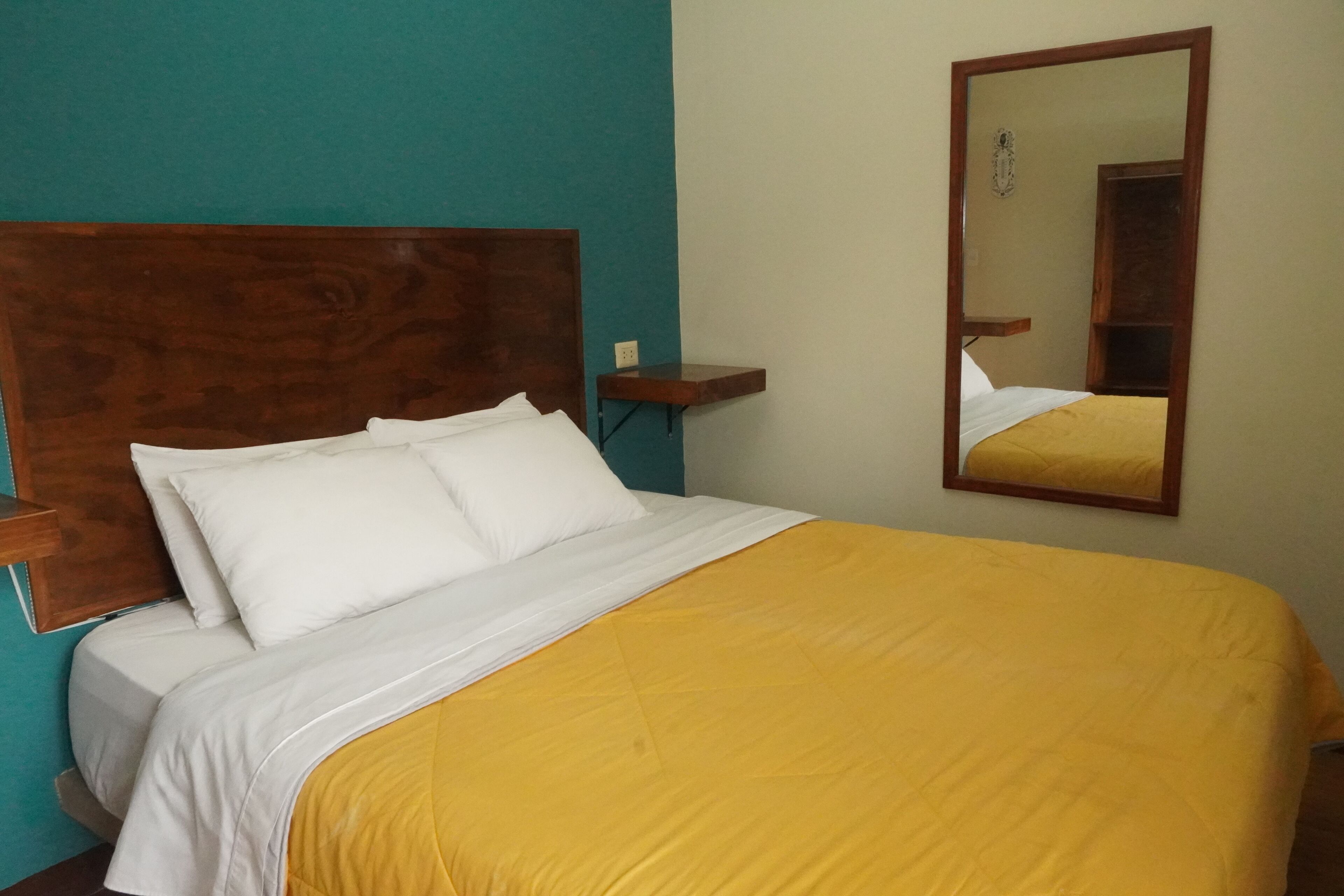 A bed with white and yellow bedding, a wooden headboard, a nightstand, and a mirror on the wall.