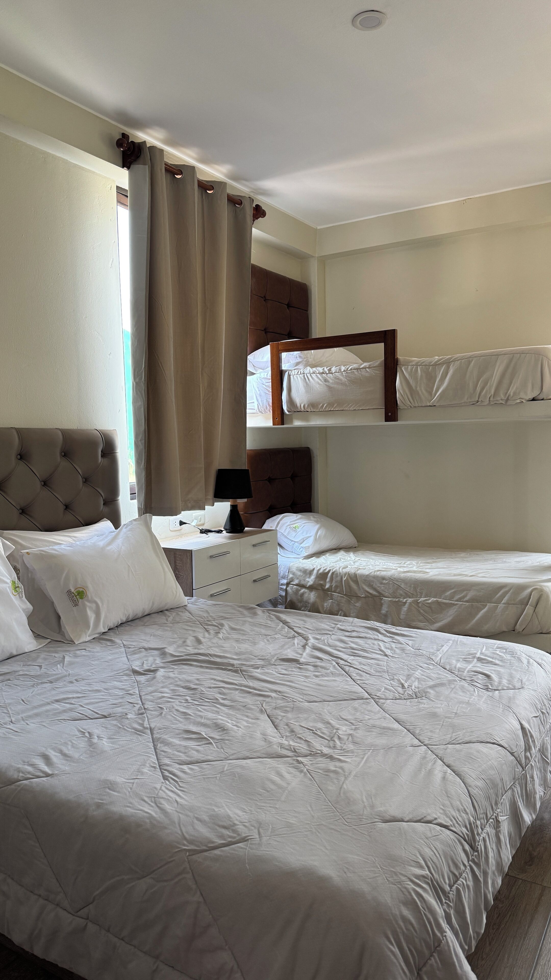 A hotel room with a bunk bed, a bedside table with a lamp, and a window with curtains.