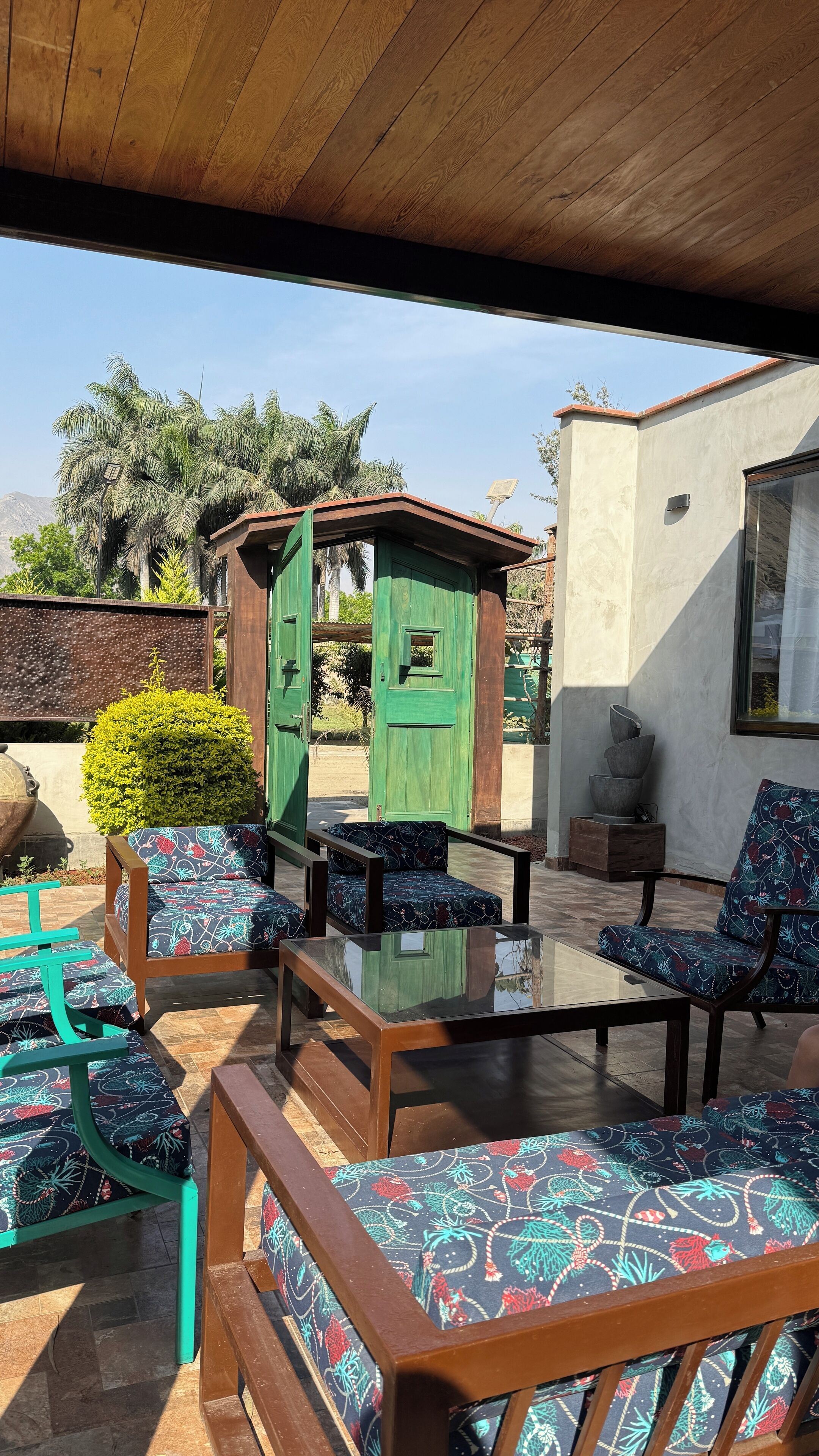 Un patio al aire libre con mobiliario de madera, incluyendo una mesa con cubierta de vidrio y sillas con cojines estampados.