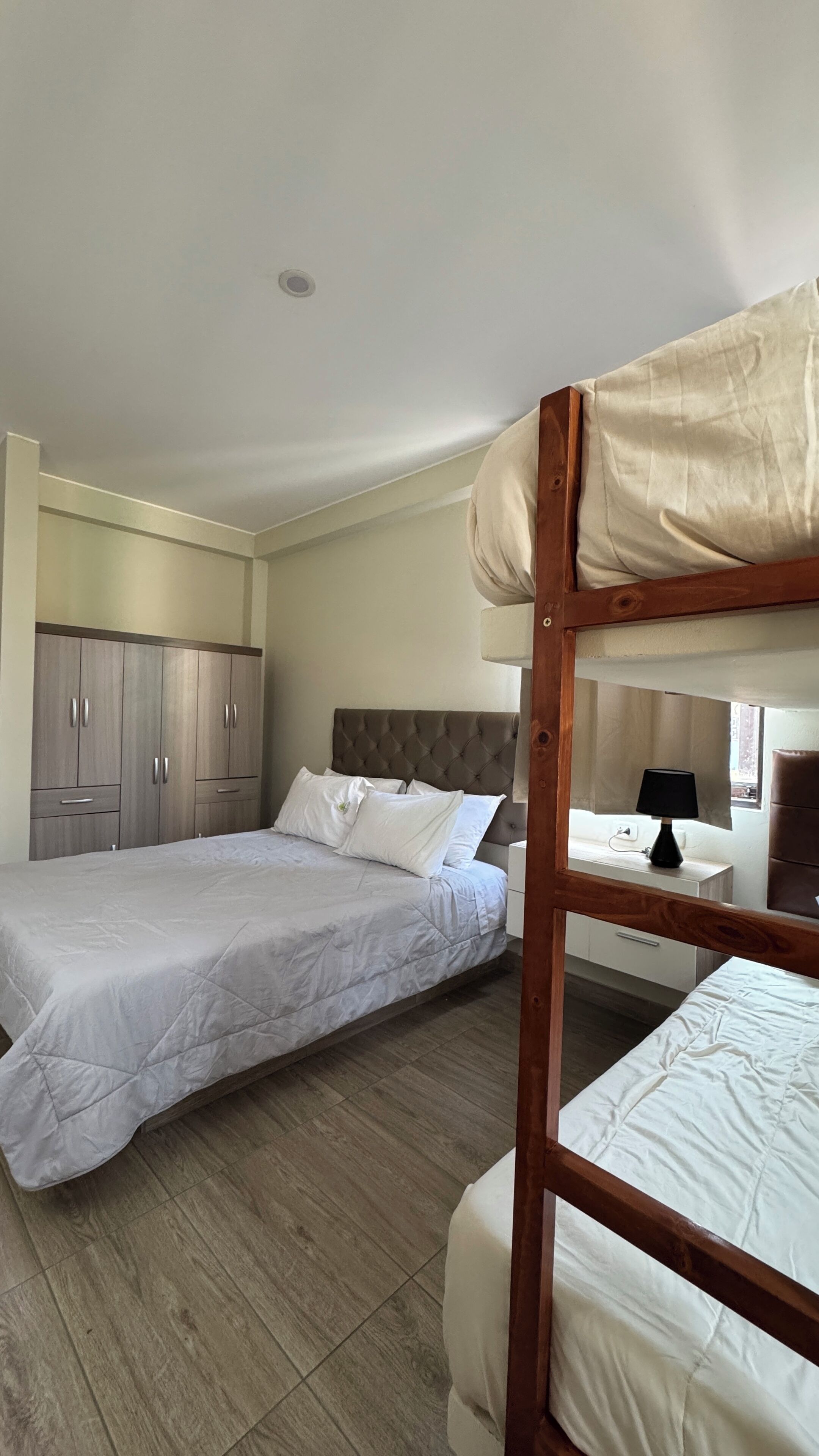 A bedroom with two beds, a wooden bunk bed, a wardrobe, and a desk with a lamp.