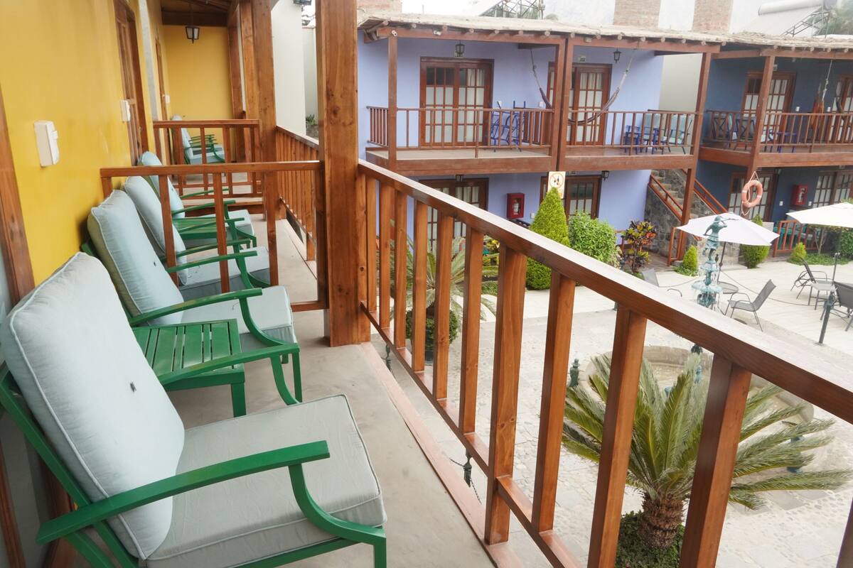 Un balcón con sillas verdes, baranda de madera y vista a un edificio con varios balcones y mesas al aire libre.