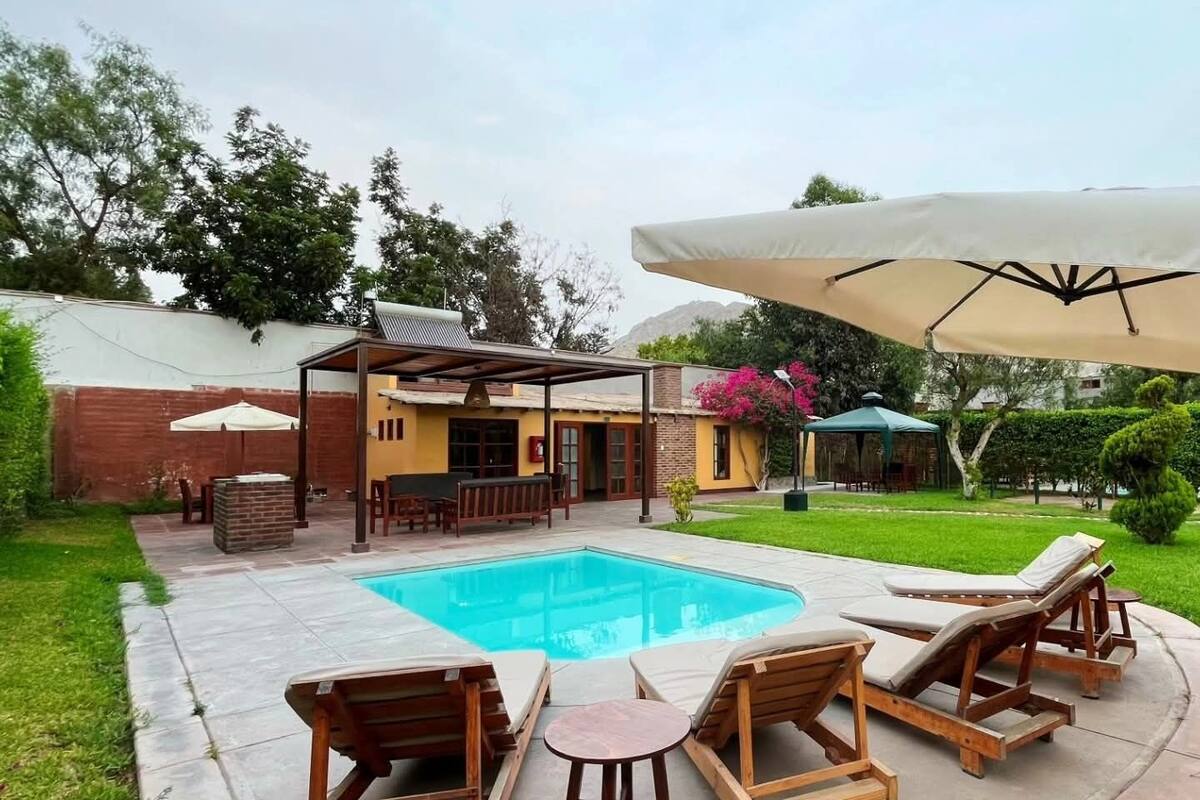 Un patio trasero con piscina, sillones de madera, un porche cubierto y un muro de ladrillo.
