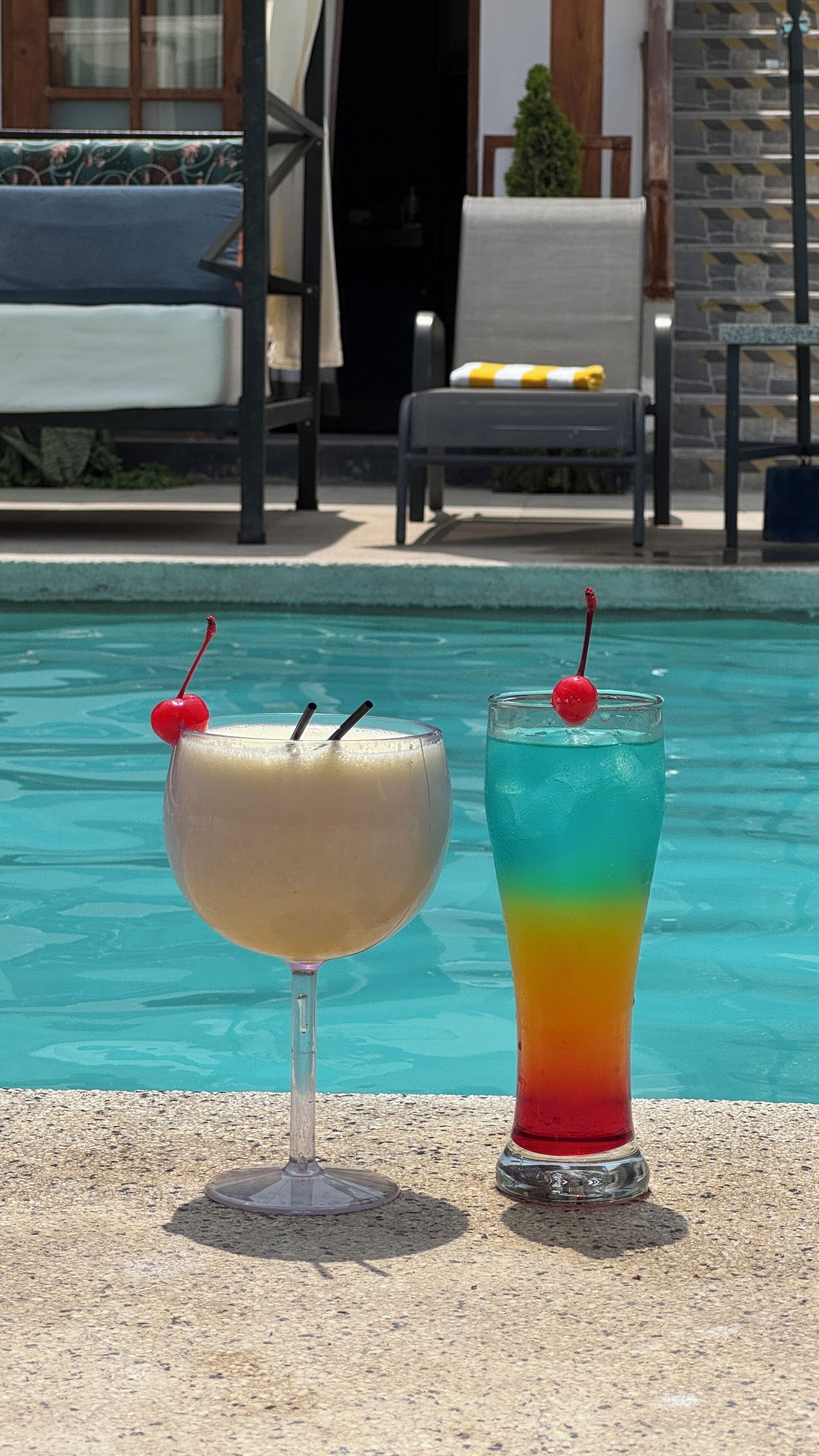 Duas bebidas perto de uma piscina com espreguiçadeiras e um edifício ao fundo.