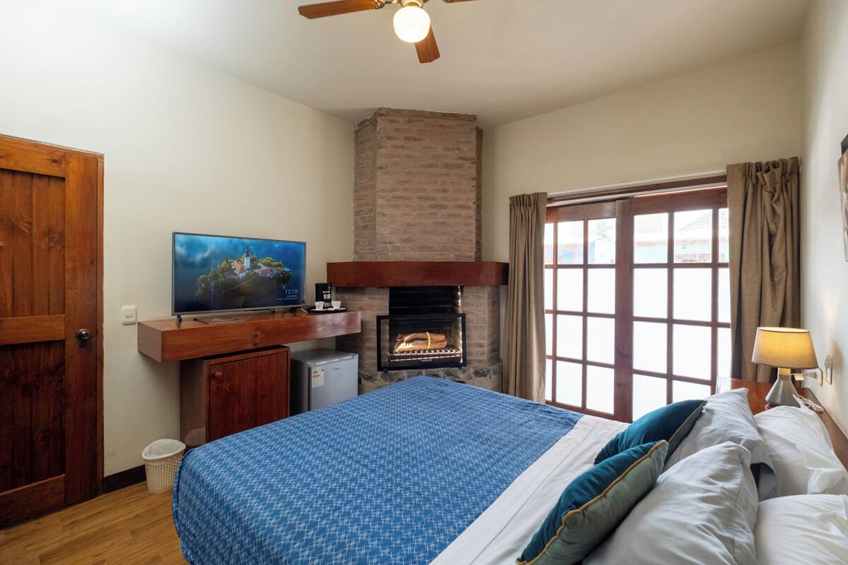 Un dormitorio con cama, televisión, chimenea, ventana y ventilador de techo.