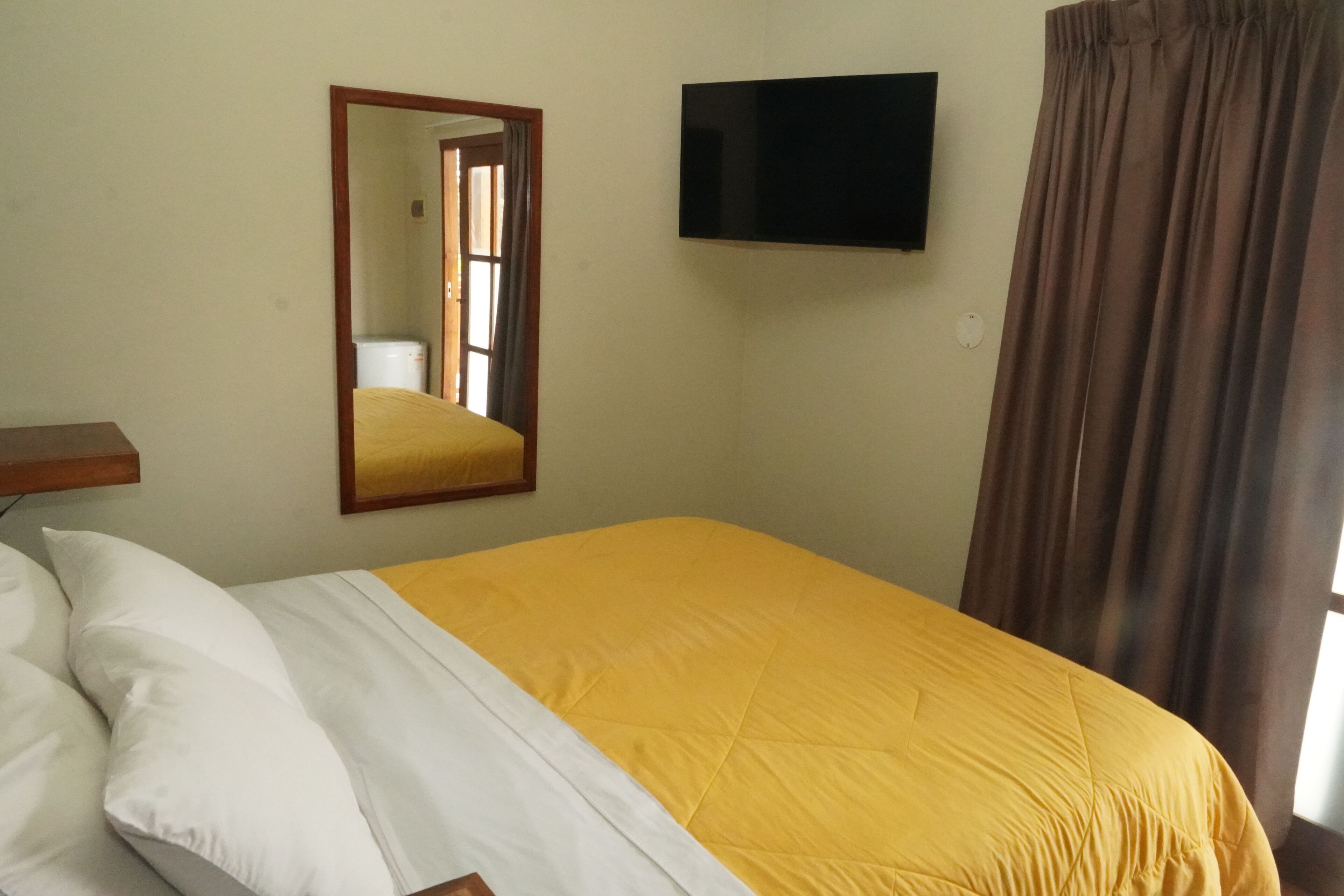 A room with a bed, a flat-screen TV mounted on the wall, a mirror, and a window with curtains.