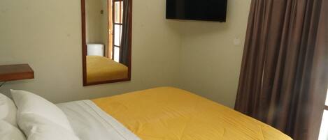 A room with a bed, a flat-screen TV mounted on the wall, a mirror, and a window with curtains.