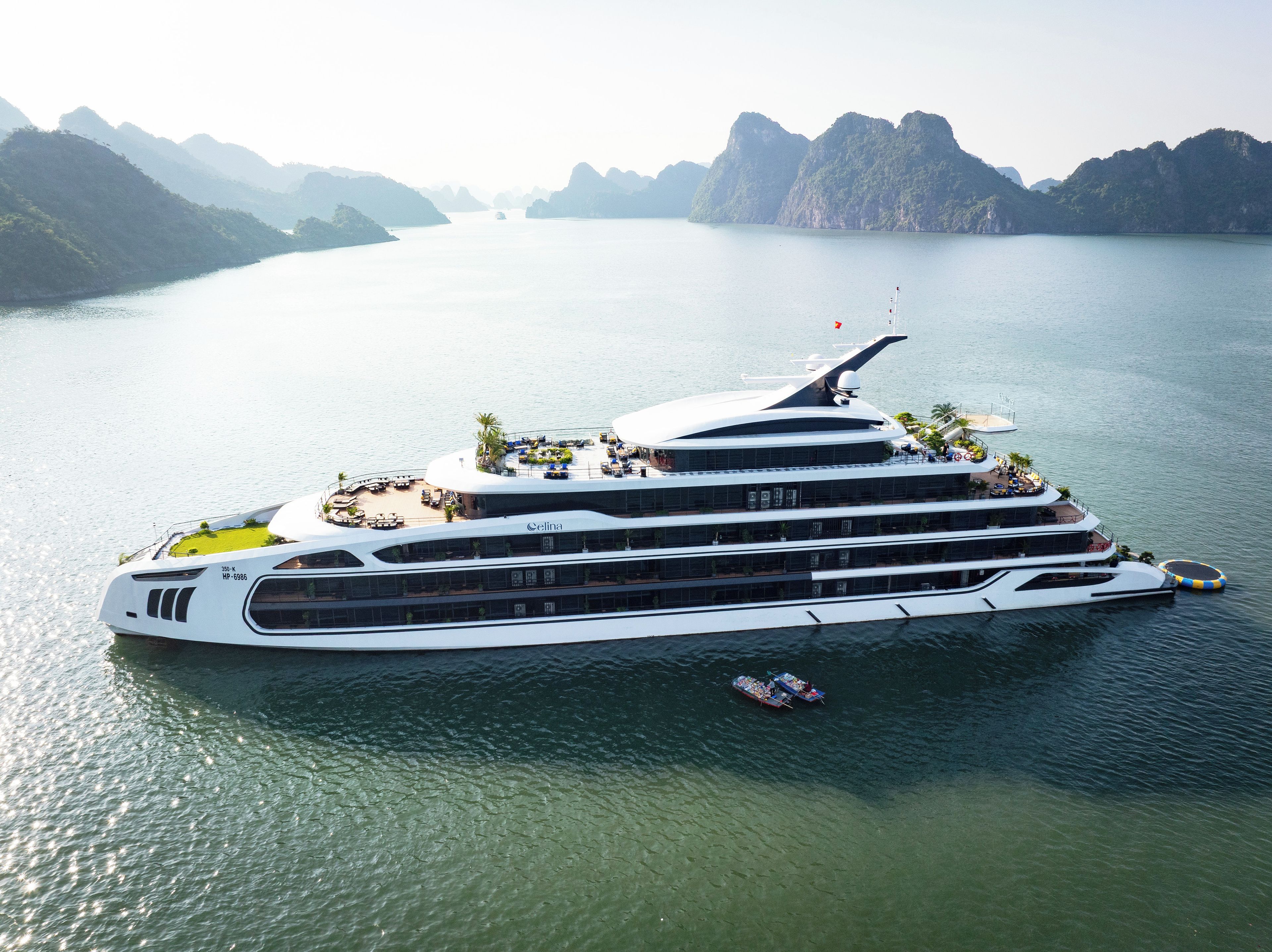 A large, multi-deck cruise ship with outdoor seating areas and a helipad, docked in a calm bay with mountainous terrain in the background.