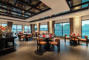 Un comedor amplio con varias mesas y sillas, ventanas grandes que ofrecen vista al agua, y un diseño de techo moderno.