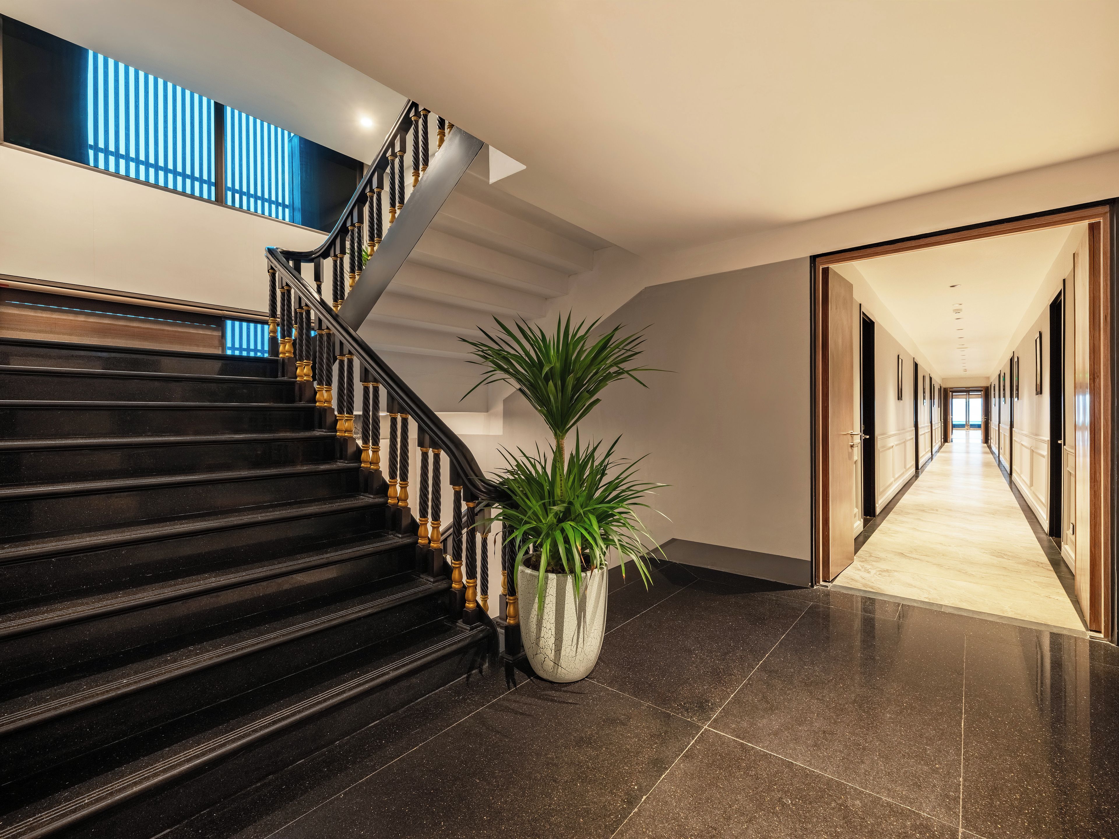 A modern hotel interior with a staircase, a potted plant, and a hallway leading to a door.