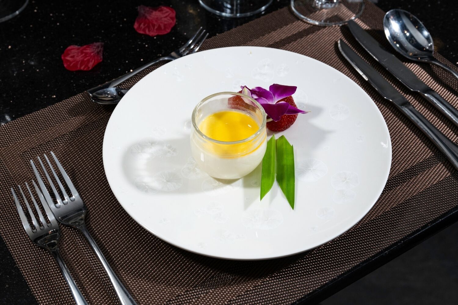 A white plate with a small glass container of yellow substance, green leaves, and a purple flower on a dark placemat.