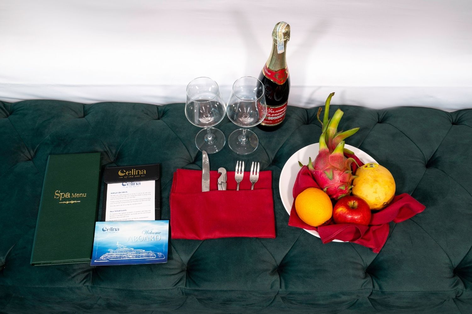 Mesa para comer con un menú, copas de vino, cubiertos, una botella de champán y un plato de fruta sobre un sofá verde con capitoné.