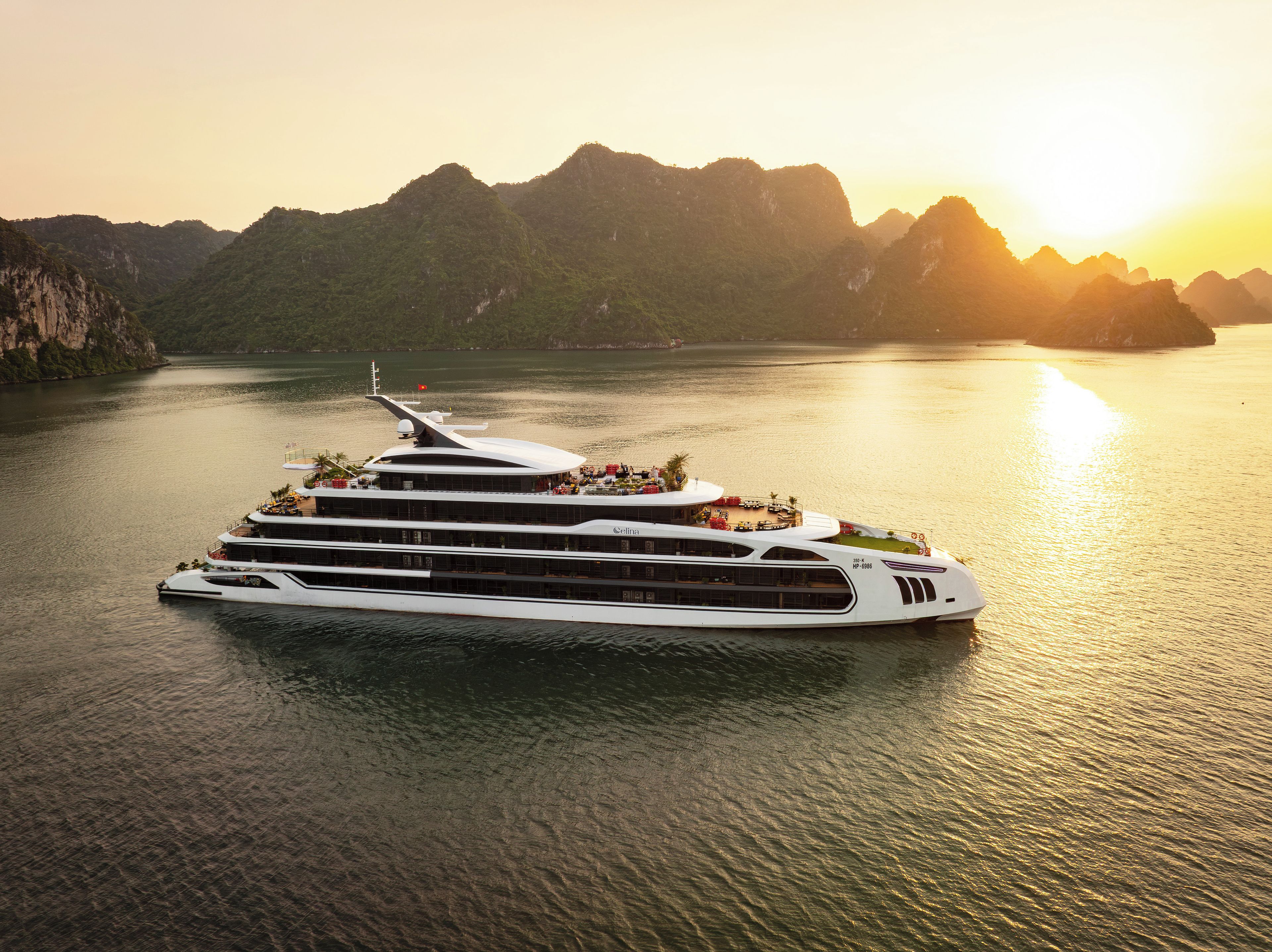 A large, multi-deck cruise ship with a modern design, docked in a calm body of water with a mountainous landscape in the background.