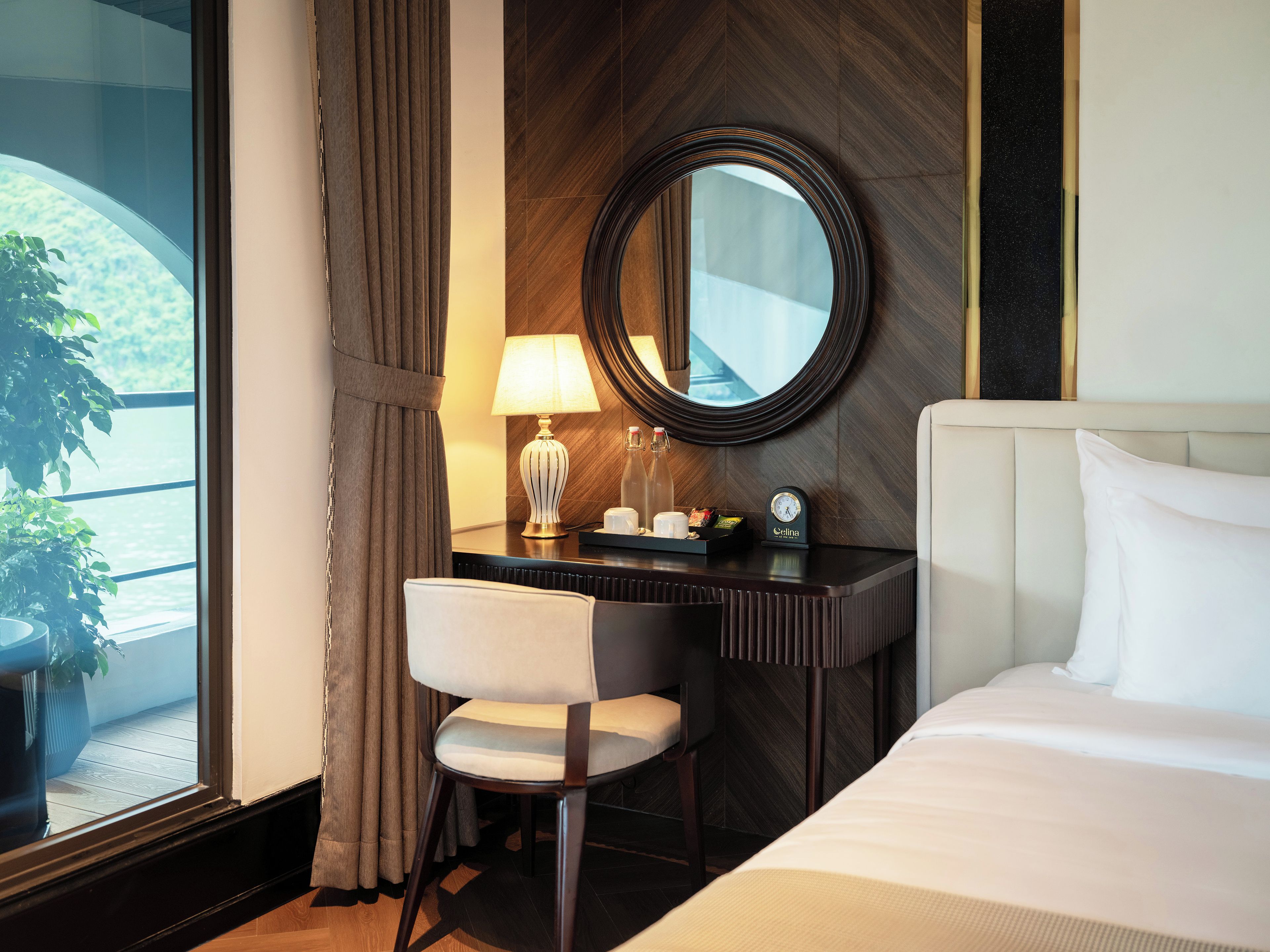 A hotel room with a bed, a desk with a lamp, a chair, a round mirror, and a view of greenery outside.