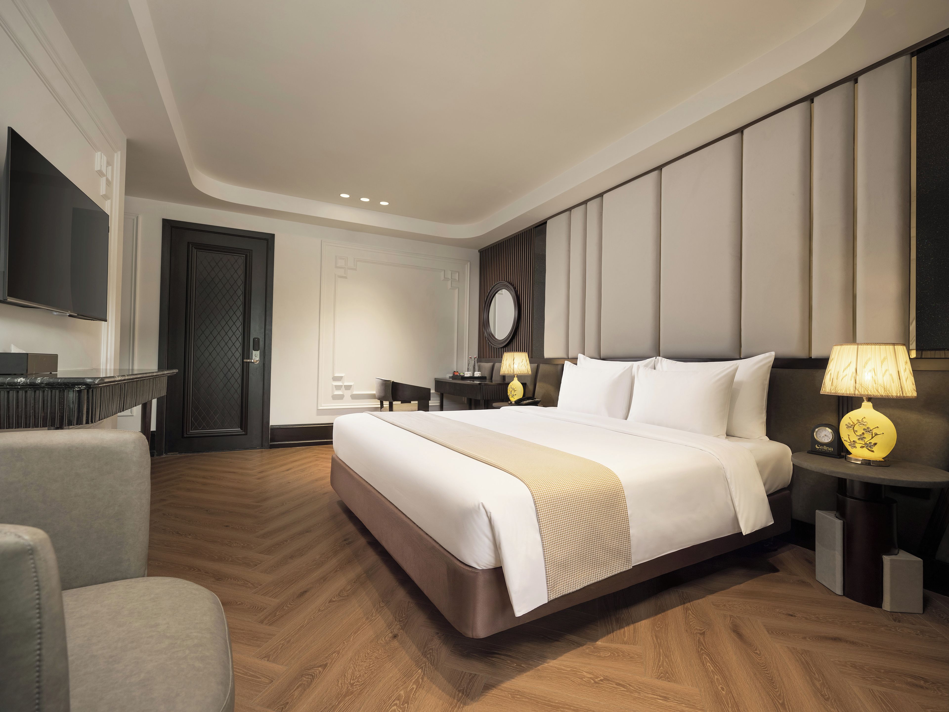 A modern hotel room with a large bed, a desk, a chair, a television, and a bedside table with a lamp.