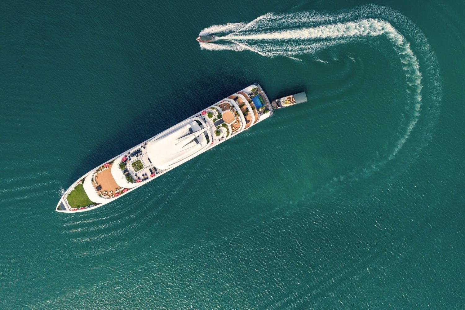 An aerial view of a cruise ship with multiple decks, a swimming pool, and a wake behind it.