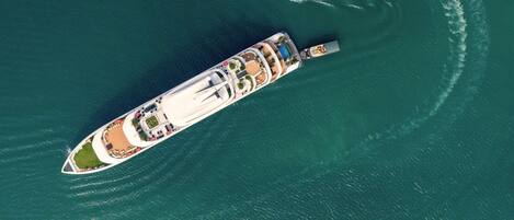 An aerial view of a cruise ship with multiple decks, a swimming pool, and a wake behind it.