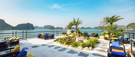 A terrace with outdoor seating, a view of the sea, and a mountainous landscape in the background.