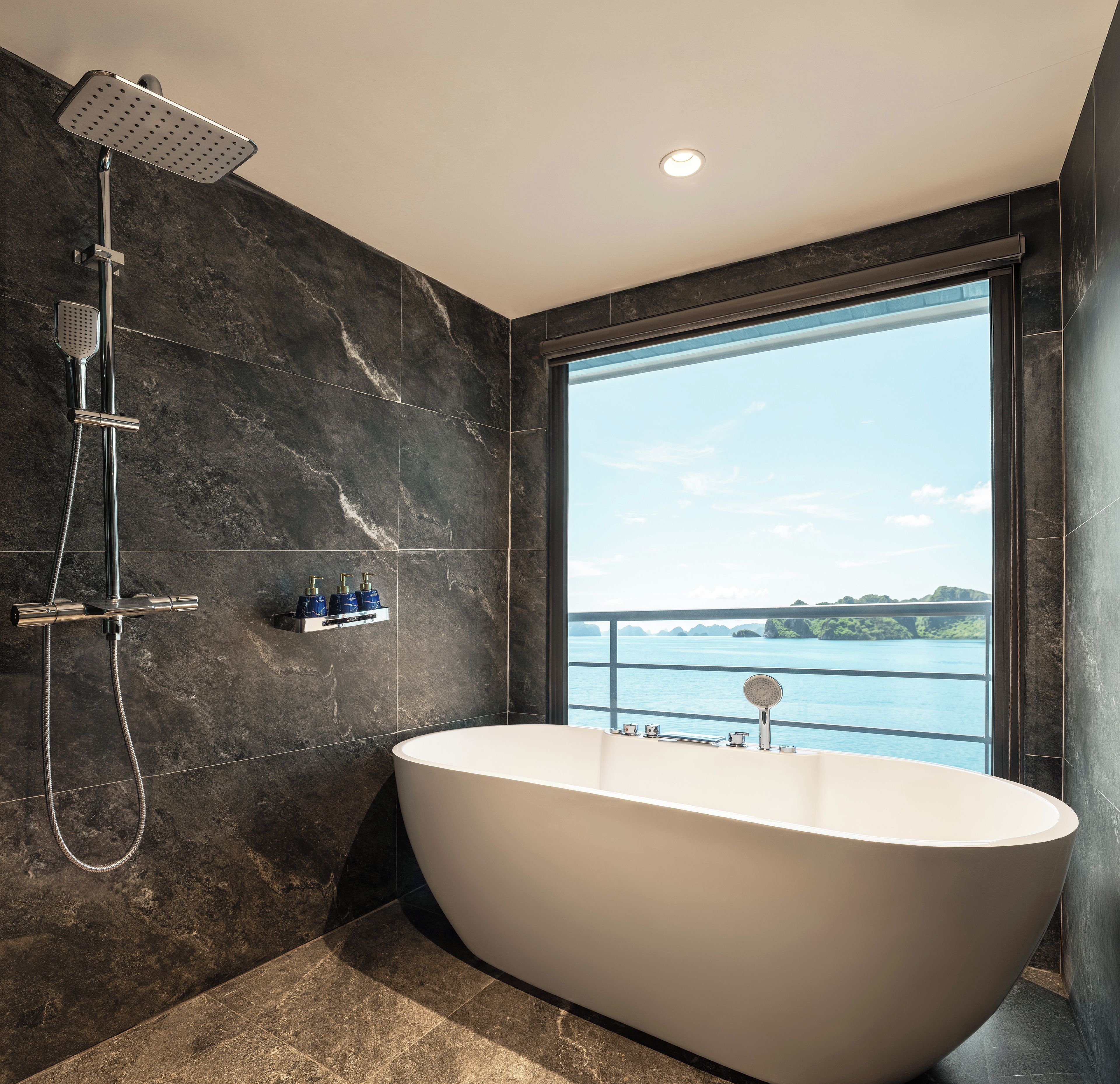 A modern bathroom with a freestanding bathtub, a showerhead, and a large window offering a view of the sea.