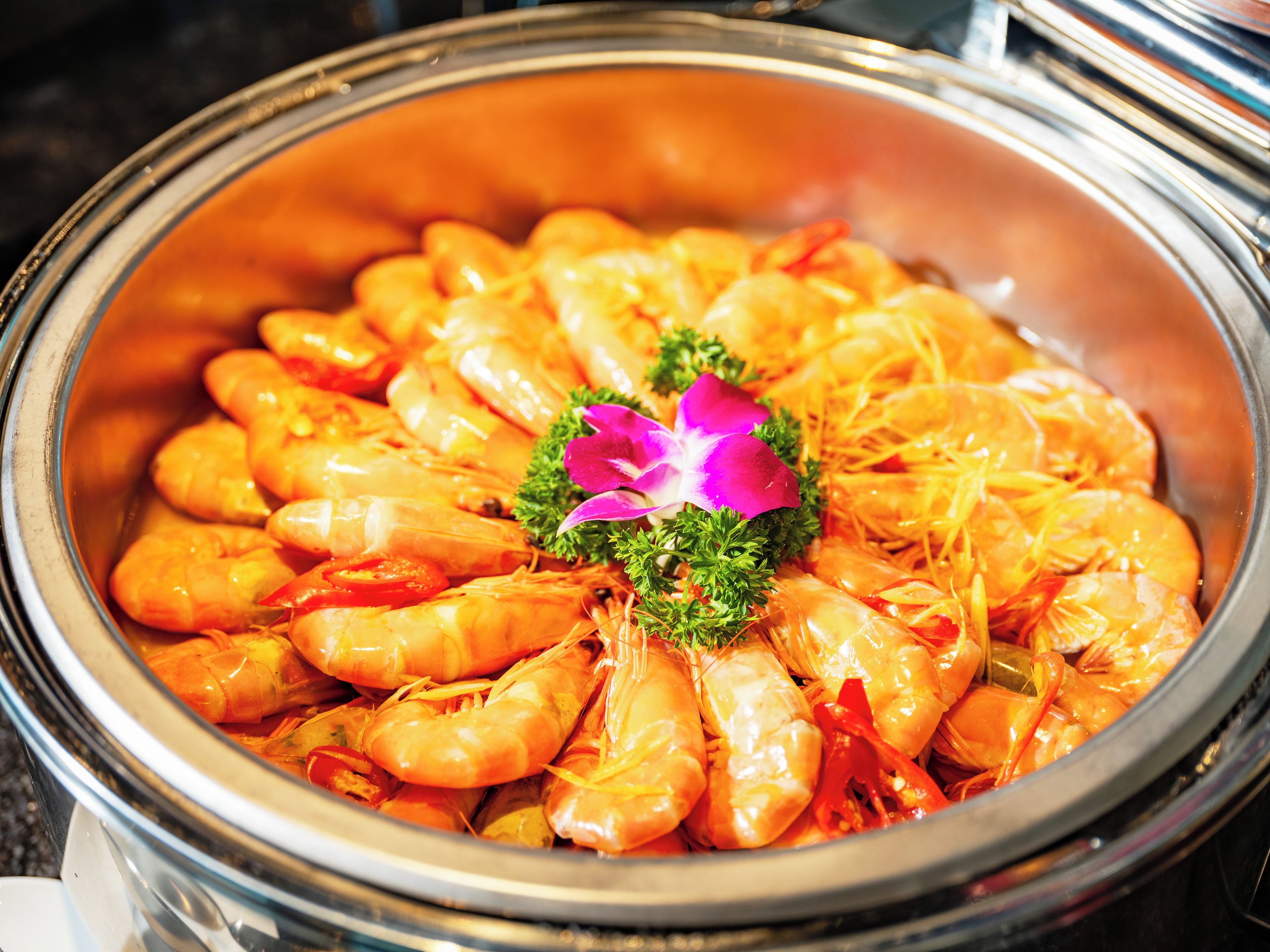 A large, round, metallic container filled with cooked shrimp, garnished with parsley and a purple flower.