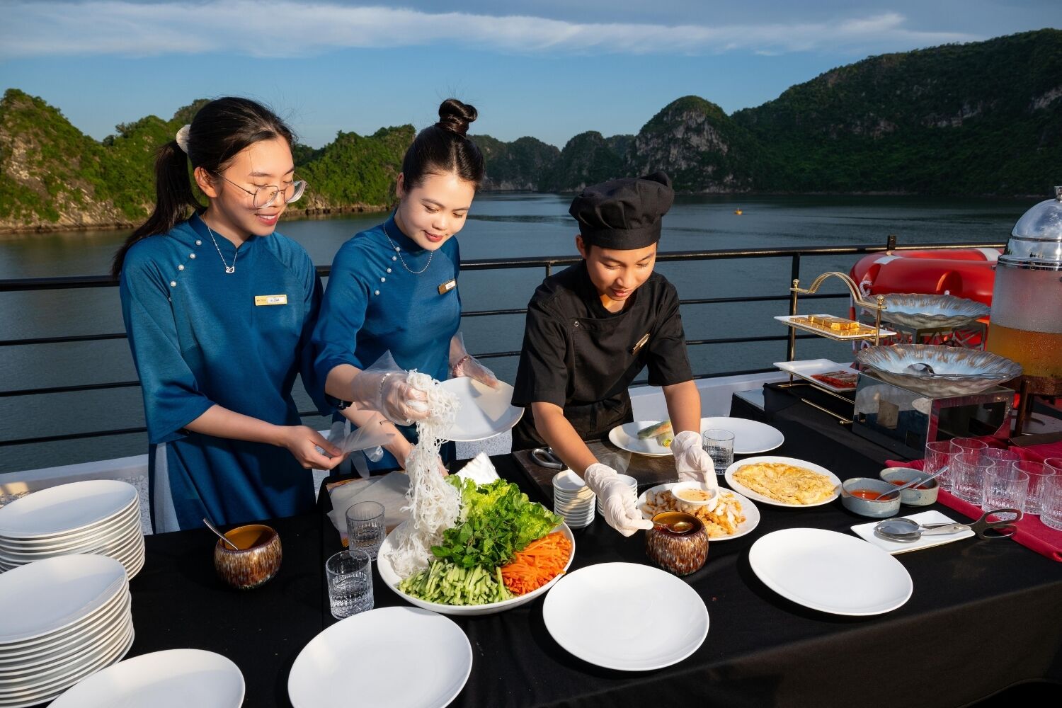 A dining table with plates, bowls, and a grill, set against a scenic lake view.