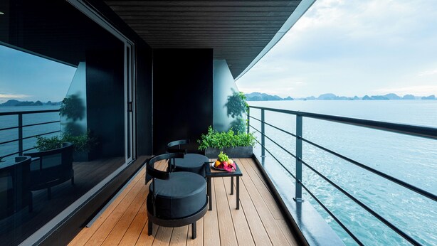 A balcony with a wooden floor, two black chairs, a small table with fruit, and a potted plant. - Celina of The Sea Cruise (Tuan Chau)