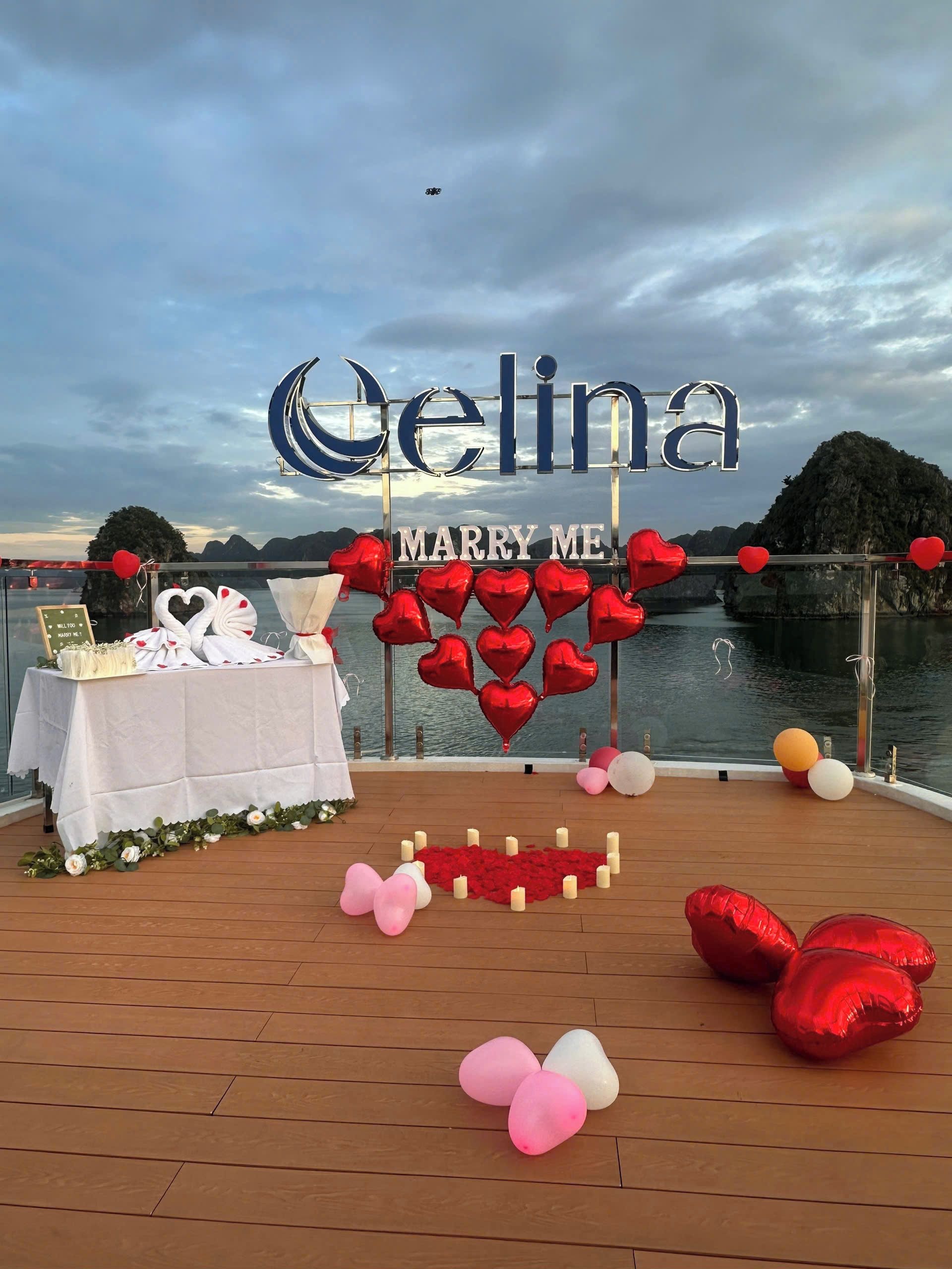 A deck with a 'MARRY ME' sign, heart-shaped balloons, and a table with a cake and flowers.