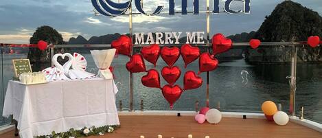 A deck with a 'MARRY ME' sign, heart-shaped balloons, and a table with a cake and flowers.