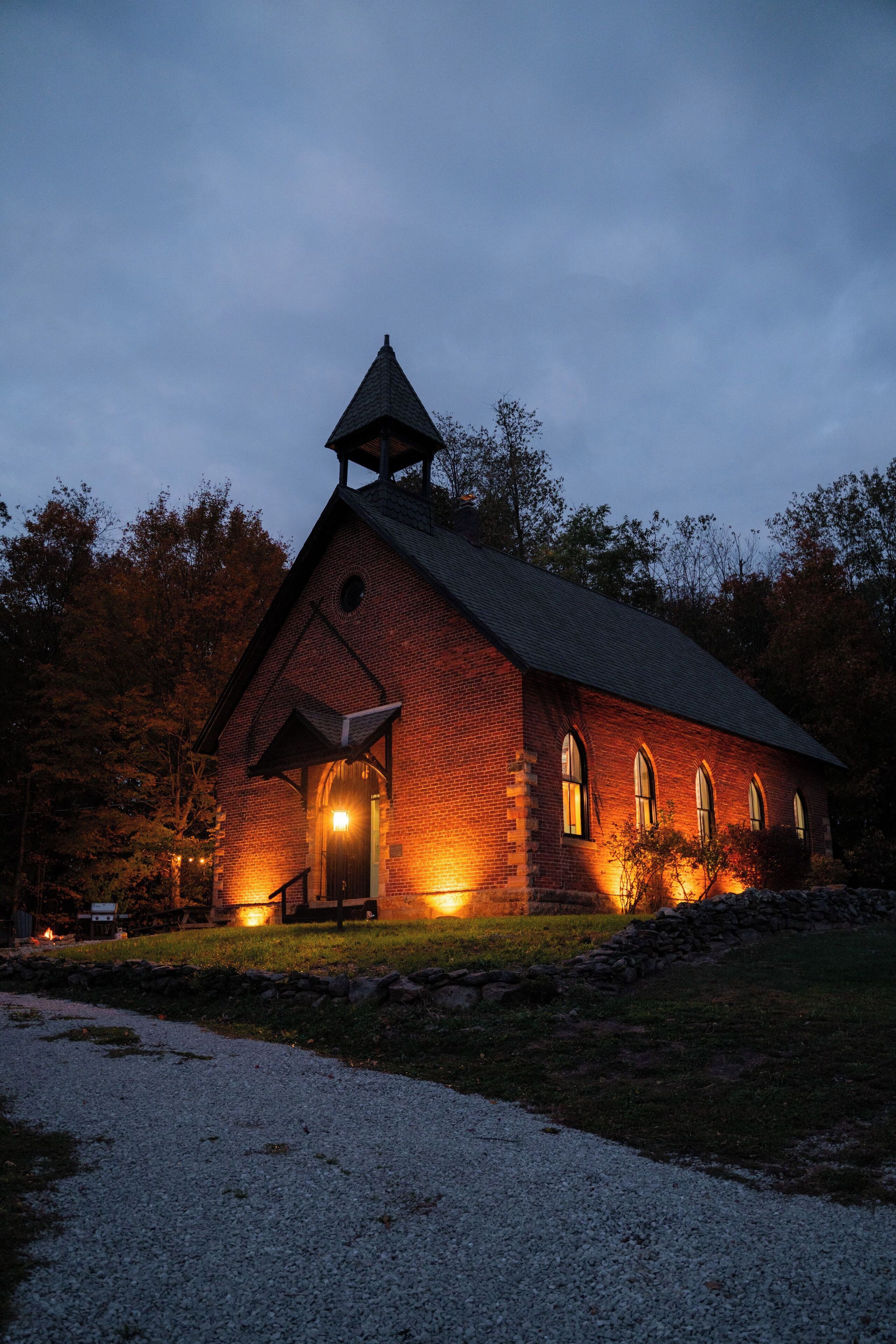 Une église en briques, dotée d’un clocher, de fenêtres illuminées et d’un chemin de gravier devant l’entrée.