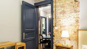Una habitación con una puerta de madera oscura, una pared de ladrillo, una mesita de noche de madera con una lámpara y un comedor al fondo.