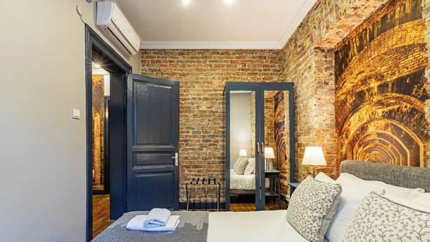 Una habitación de hotel con pared de ladrillo, una cama con ropa de cama estampada, una mesita de noche con lámpara y un espejo.