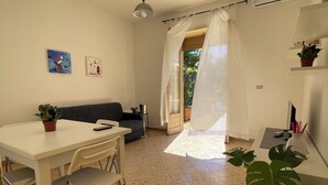 Una habitación con una mesa de comedor, sillas, un sofá, una pequeña planta en maceta y una planta grande en maceta.