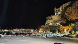 Puerto deportivo con barcos amarrados, un edificio grande con una torre en un acantilado y una zona ribereña bien iluminada.