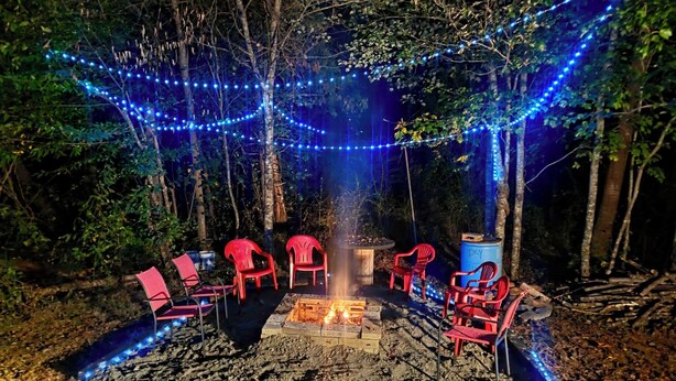 Un espacio de estar al aire libre con sillas de plástico rojas dispuestas alrededor de un brasero central, rodeado de una zona boscosa con luces azules colgantes.