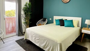 Une chambre à coucher avec un lit, une horloge au mur, un ventilateur, une table de chevet et une vue sur l’extérieur.