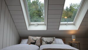 Een slaapkamer met twee dakramen, een bed met wit beddengoed, twee kussens met hertenmotieven en een nachtkastje met een lamp.