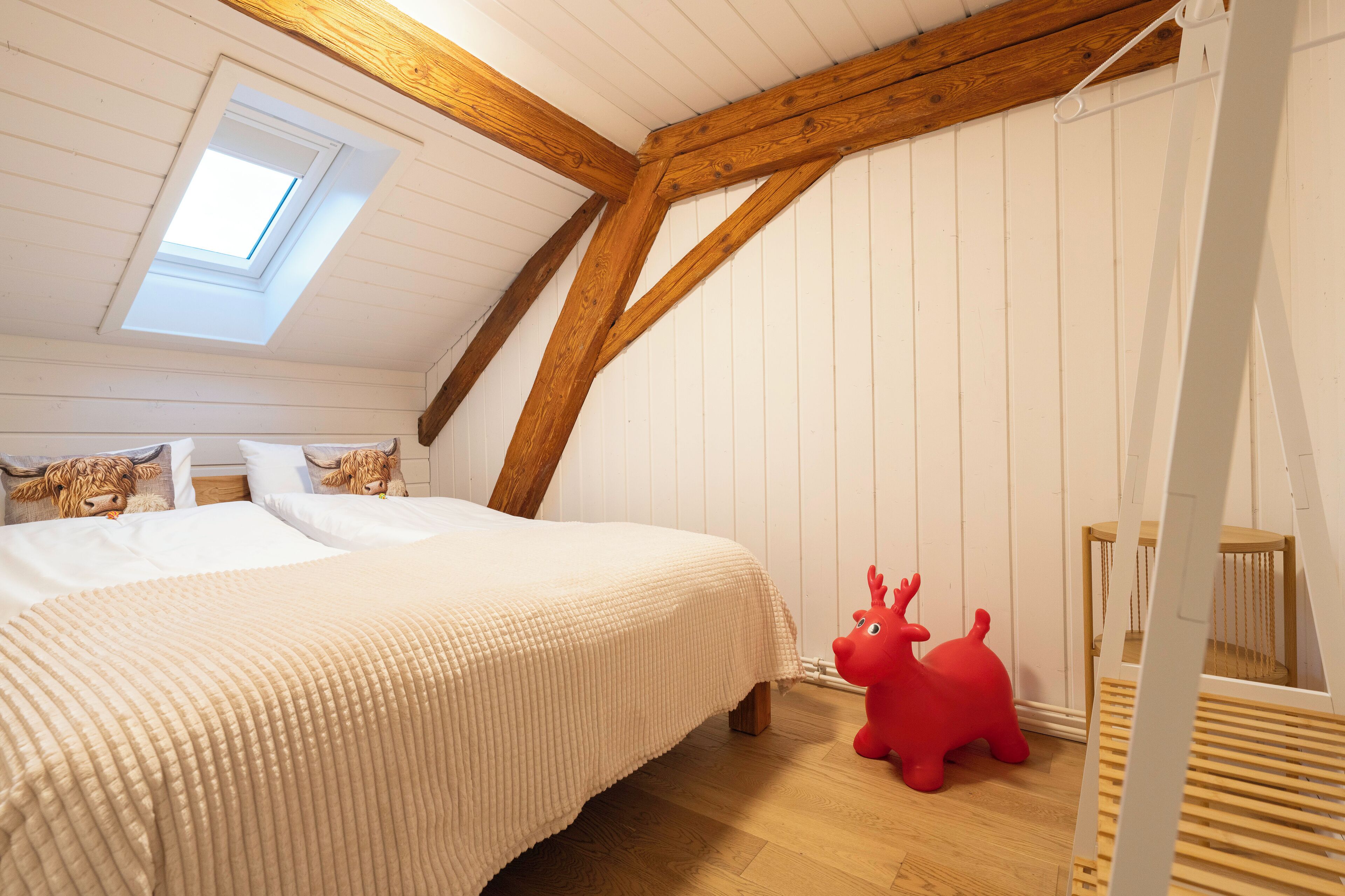 Une chambre avec un plafond en pente, un puits de lumière, un lit recouvert d’une couverture beige, un jouet rouge et un plancher en bois.