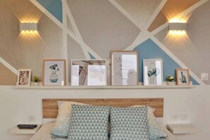 A bedroom with a wooden headboard, a bed with patterned pillows, a bedside table, and a wall-mounted shelf with framed pictures and a small plant.